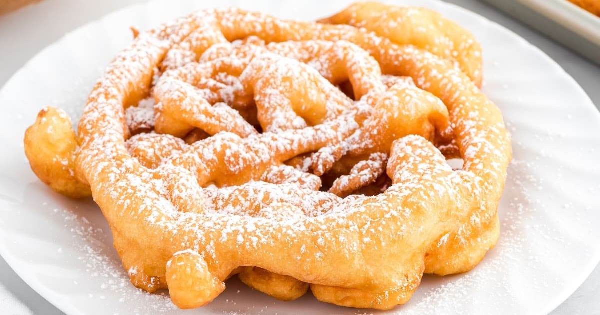 Classic funnel cake covered in powdered sugar on a white plate, shown up close with golden crispy edges.