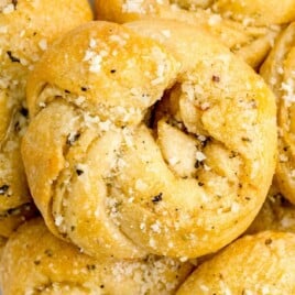 Golden garlic knots topped with herbs and shredded Parmesan on a white plate.