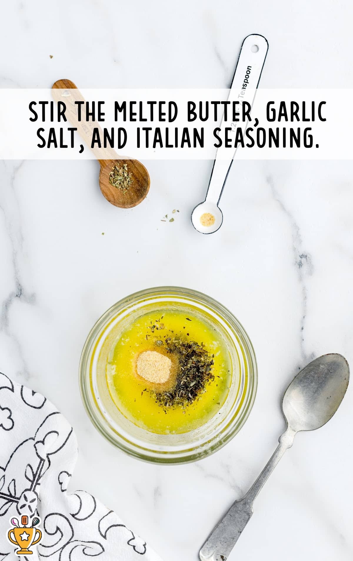 Small bowl of melted butter with garlic salt and Italian seasoning on a white counter with measuring spoons.