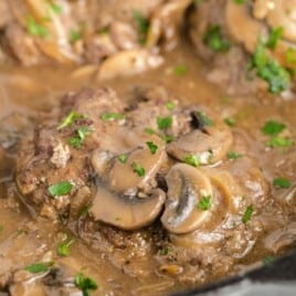 Close-up of hamburger steaks simmering in a skillet full of savory mushroom and onion gravy, topped with fresh parsley.