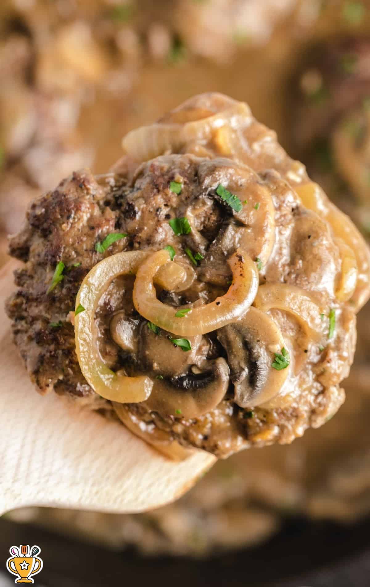 Close-up of a juicy hamburger steak covered in onion and mushroom gravy, being lifted from a skillet with a wooden spoon.