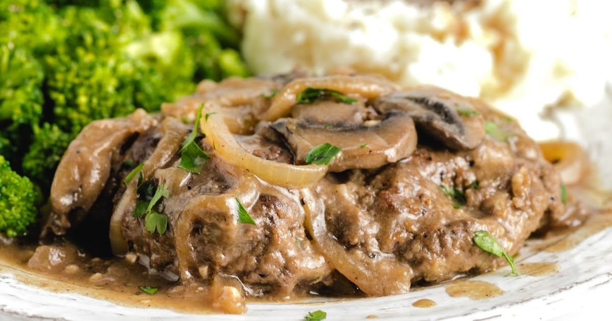 Plate of hamburger steak with mushroom gravy served alongside steamed broccoli and creamy mashed potatoes topped with brown gravy.
