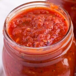 Full jar of homemade tomato sauce with a spoonful on top, rich in tomatoes and seasonings, with blurred background.