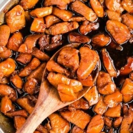 Wooden spoon lifting glazed honey garlic chicken from a skillet filled with saucy, bite-sized pieces.