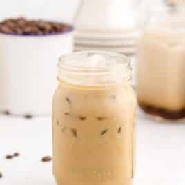 Straight-on shot of a mason jar filled with iced caramel macchiato, with coffee beans and other drink jars blurred in the background.