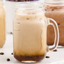 Four mason jars filled with various shades of iced caramel macchiato lined up on a counter with coffee beans scattered around.
