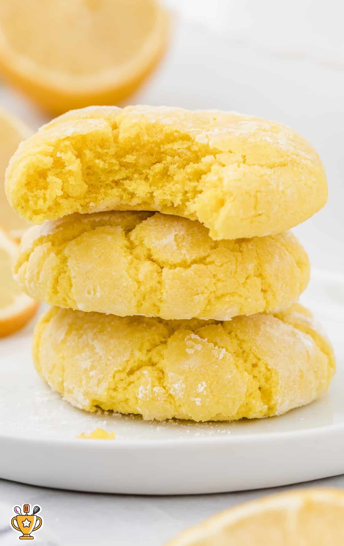 Stack of three lemon crinkle cookies with a bite taken from the top, sitting on a white plate with lemon slices in the background.