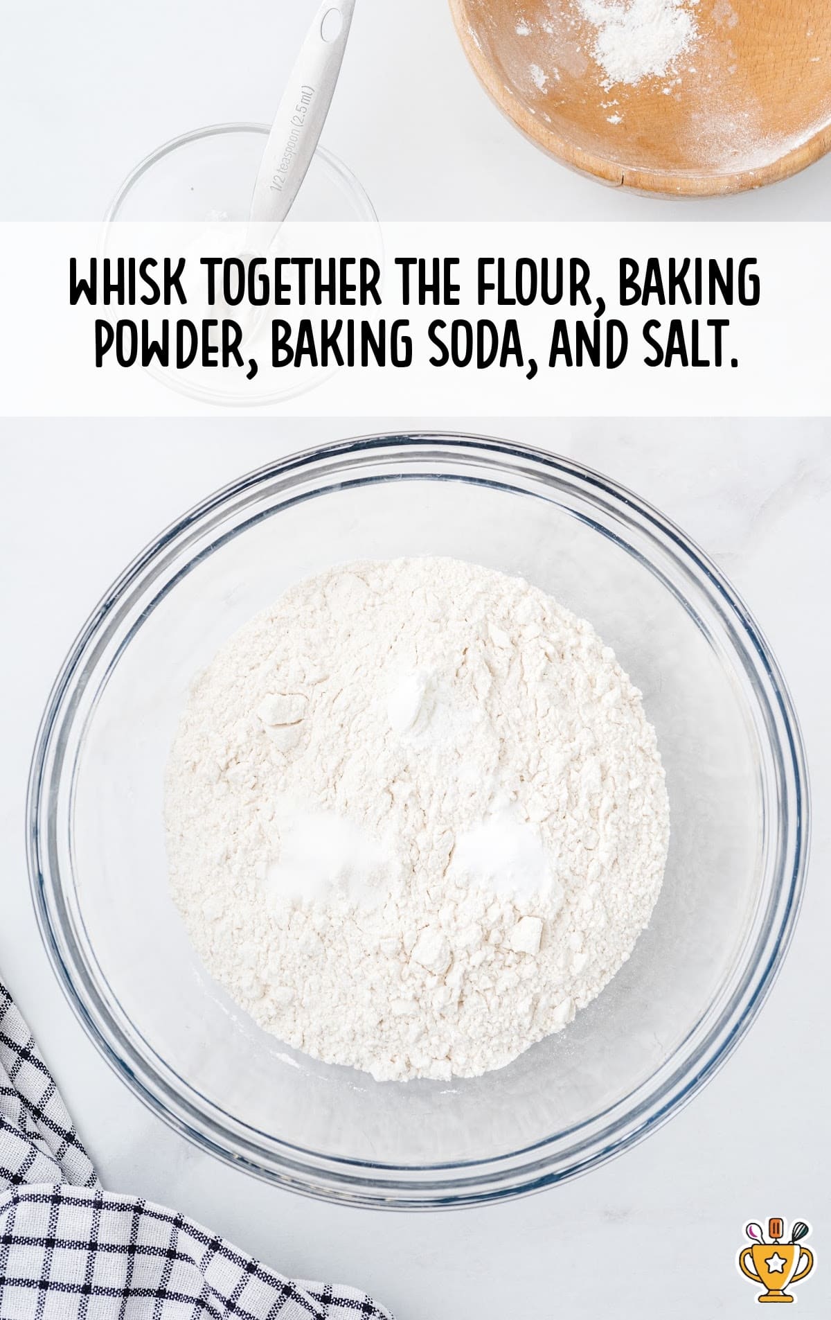 Clear bowl with flour, baking powder, baking soda, and salt, with a measuring spoon and a checked towel on the side.