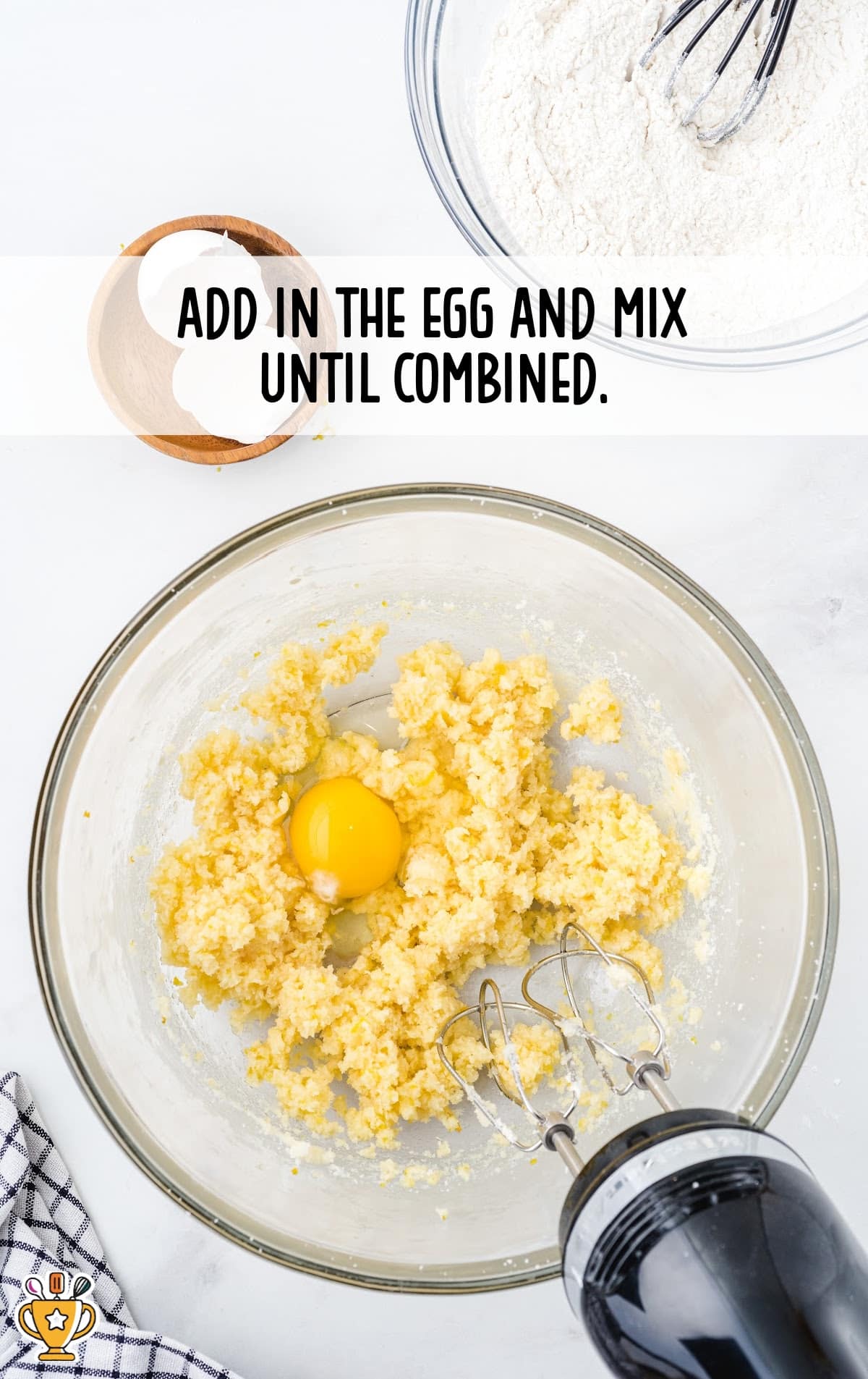 Egg added to creamed butter and sugar mixture, sitting in a mixing bowl with beaters, flour bowl in the background.
