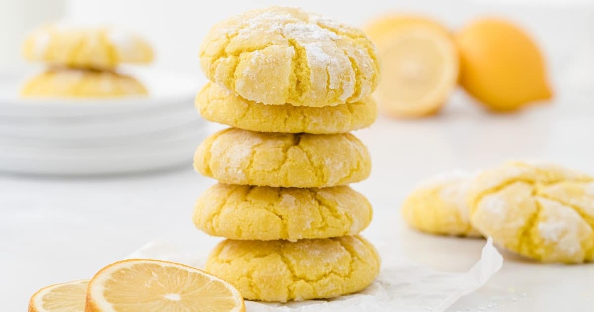 Five lemon crinkle cookies stacked in a tower, with lemons and more cookies in the background and a lemon slice in the foreground.