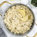 Top-down view of a large pot of cooked lemon rice with two lemon wedges in the center, surrounded by marble counter, fresh thyme, and kitchen towel.