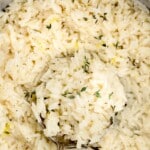 Overhead shot of a pan filled with lemon rice garnished with thyme and lemon zest, a vintage spoon scooping from the center, and sliced lemons and a small bowl of herbs in the background.