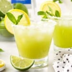 Two mint margaritas served in clear glasses with salted rims, lemon and mint garnish, and fresh limes scattered in the background.