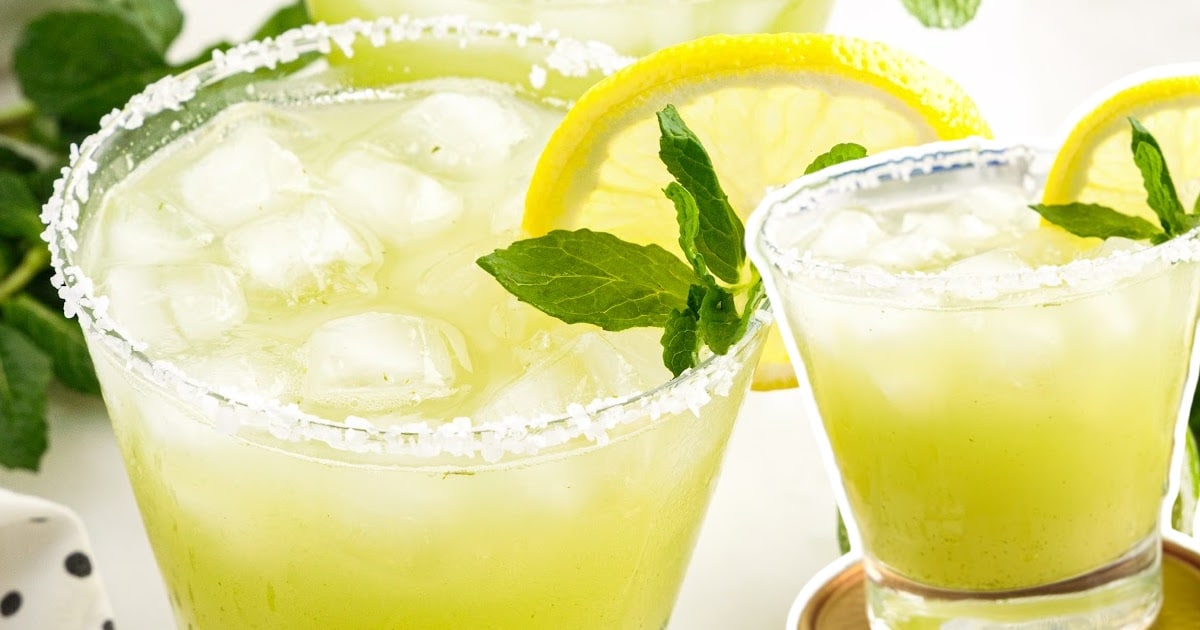 Two mint margaritas in focus with lemon and mint garnish, one in front of a bottle of tequila, lime wedge in foreground.