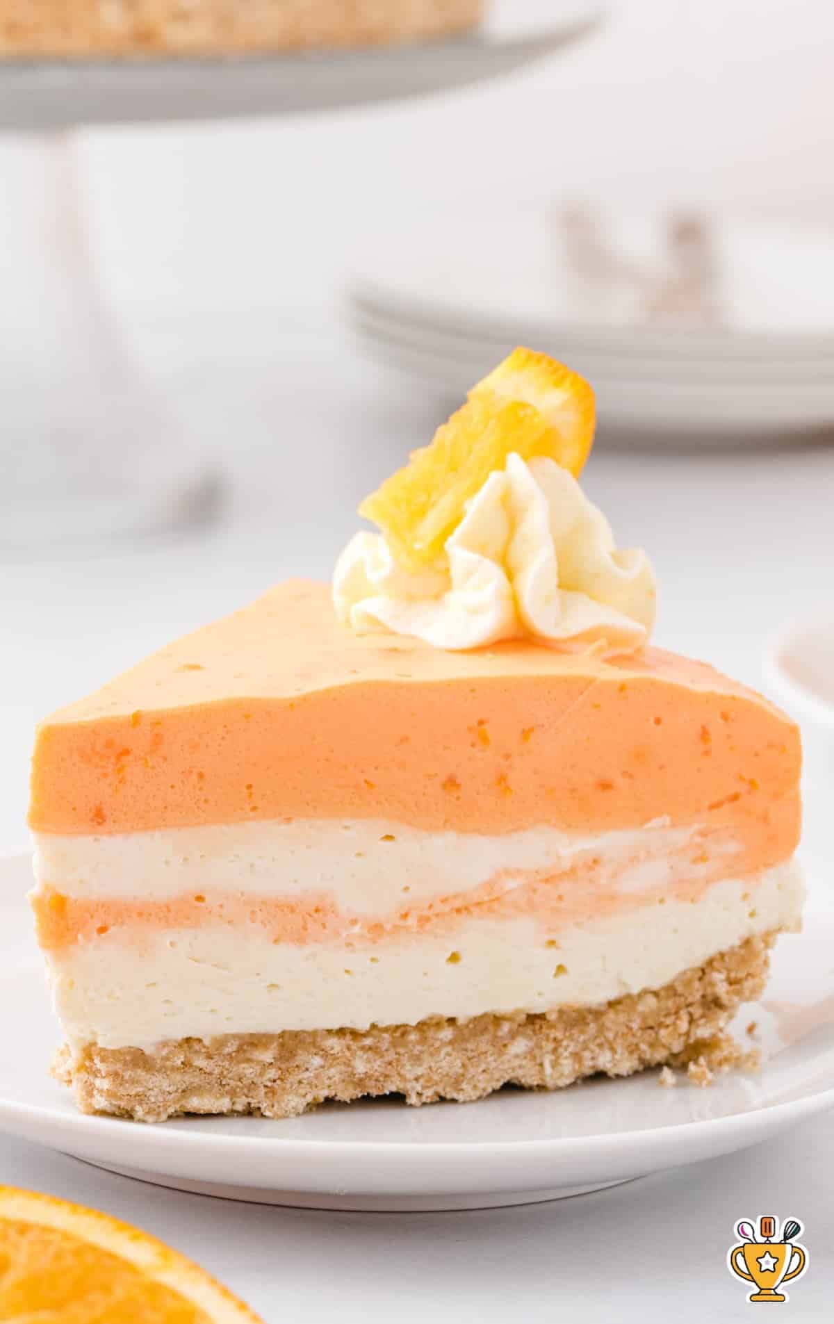 A front-facing view of a single slice of layered orange creamsicle cheesecake on a plate, topped with a swirl of whipped cream and orange garnish.