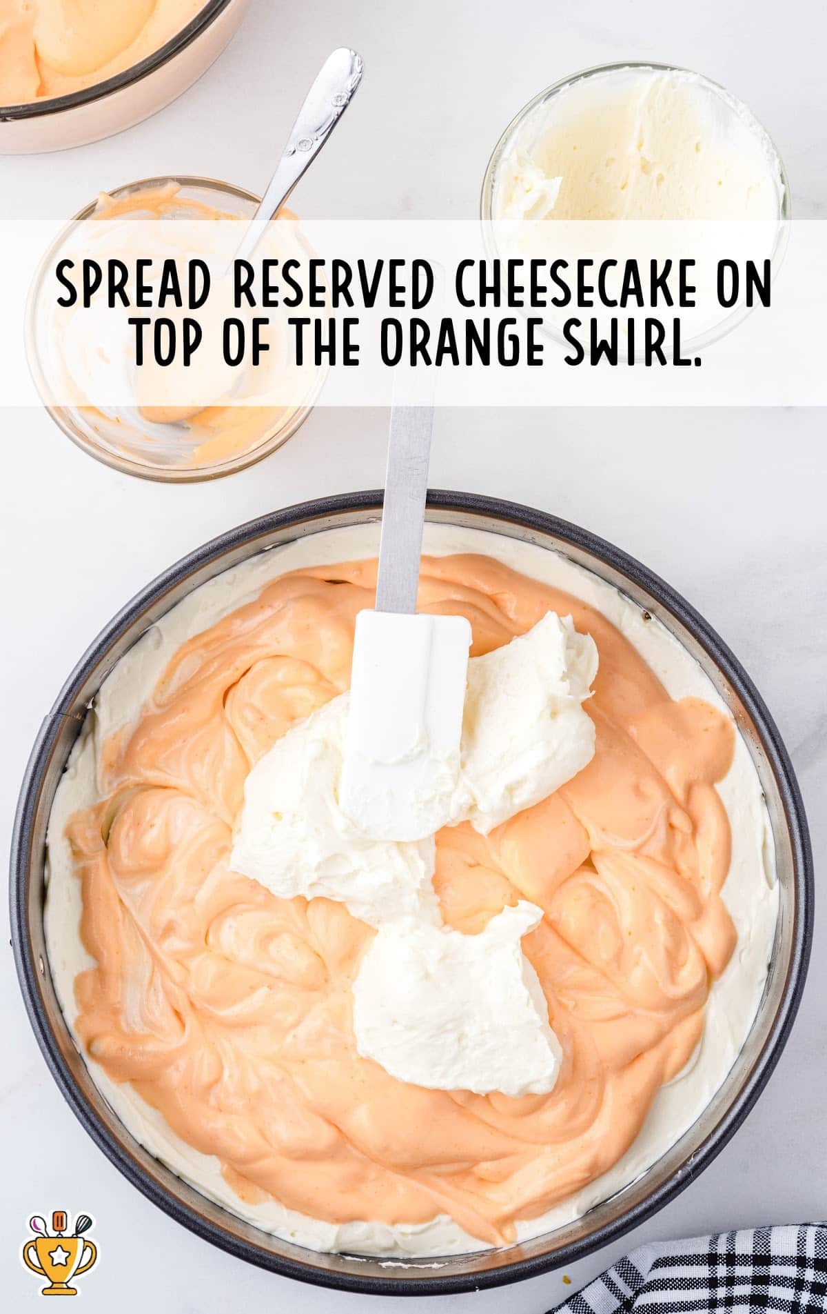 Springform pan with orange swirl mixture partially spread over a cheesecake layer, being topped with reserved white cheesecake mixture using a spatula.