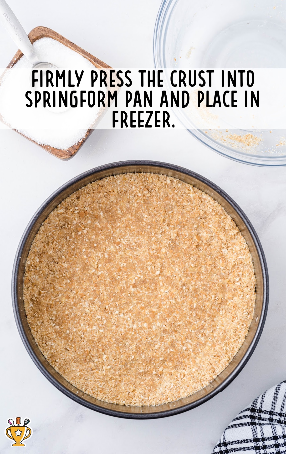 Close-up view of a springform pan filled with tightly packed vanilla wafer crust, sitting on a white surface with sugar nearby.