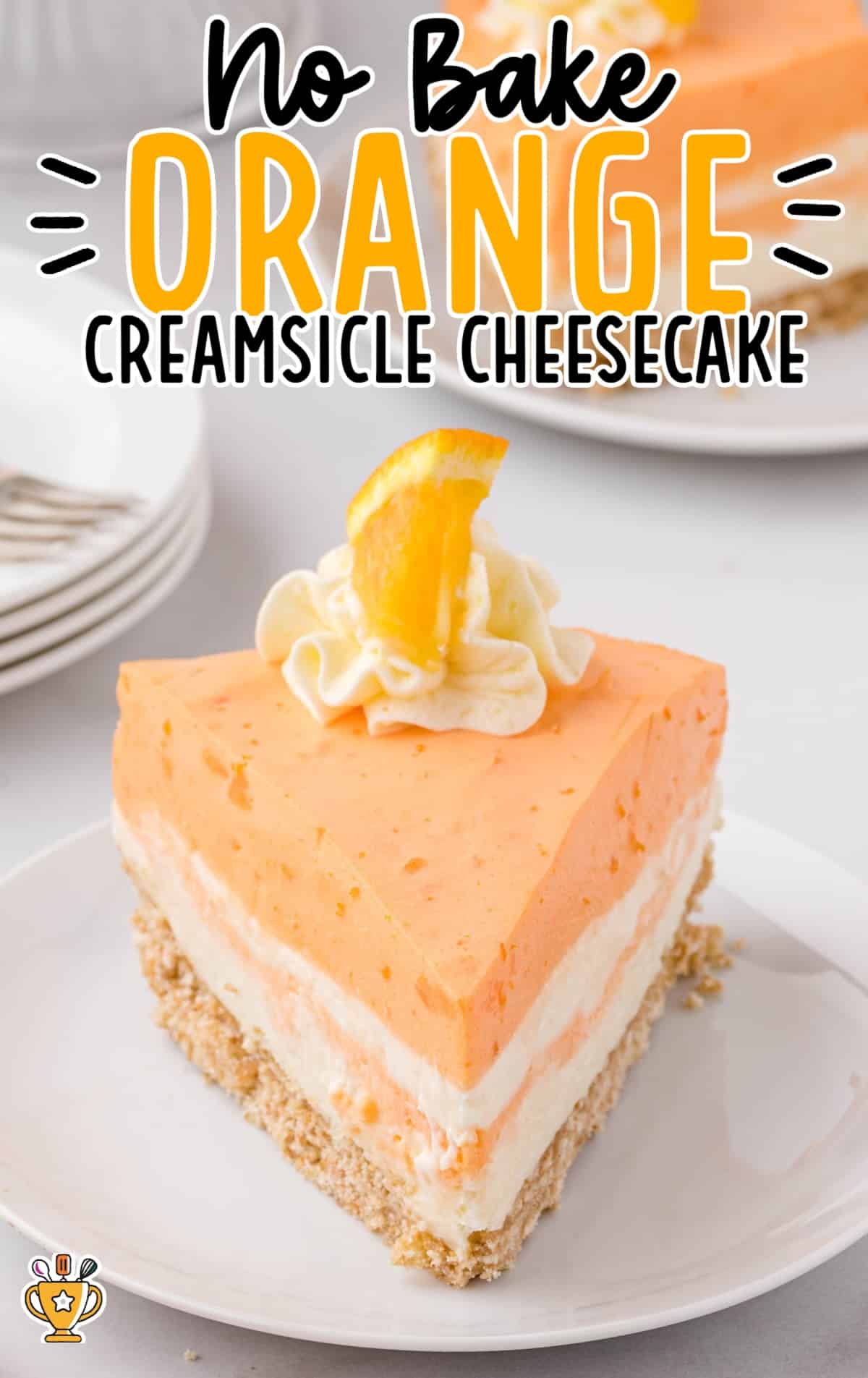 A tall slice of no-bake orange creamsicle cheesecake topped with whipped cream and a wedge of orange, plated with stacked dishes in the background.