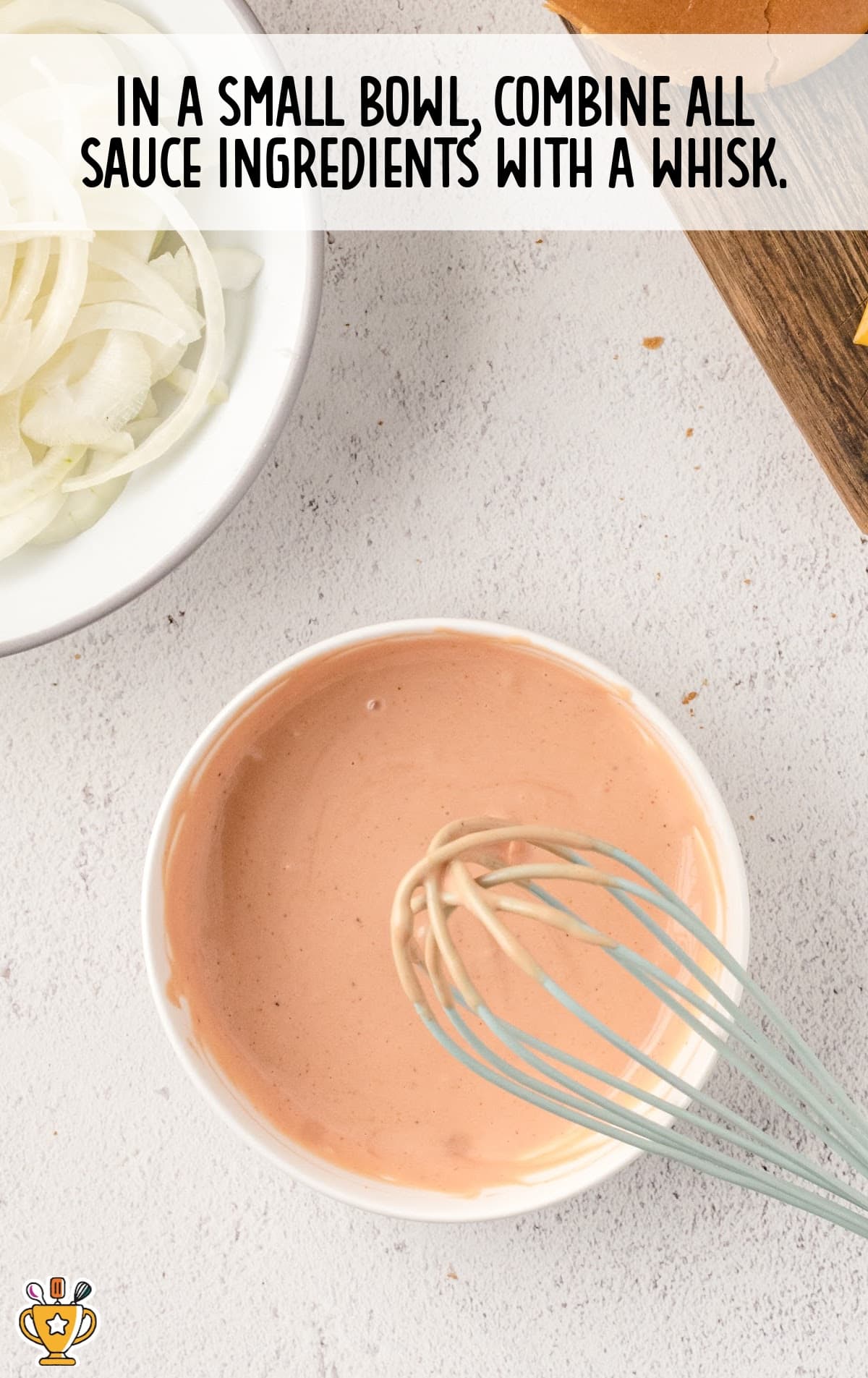 Overhead view of a bowl of burger sauce with a whisk, surrounded by sliced onions and sandwich bun ingredients.