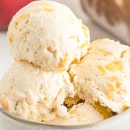 Bowl filled with scoops of peach ice cream in focus, with fresh peaches and ice cream container in background.