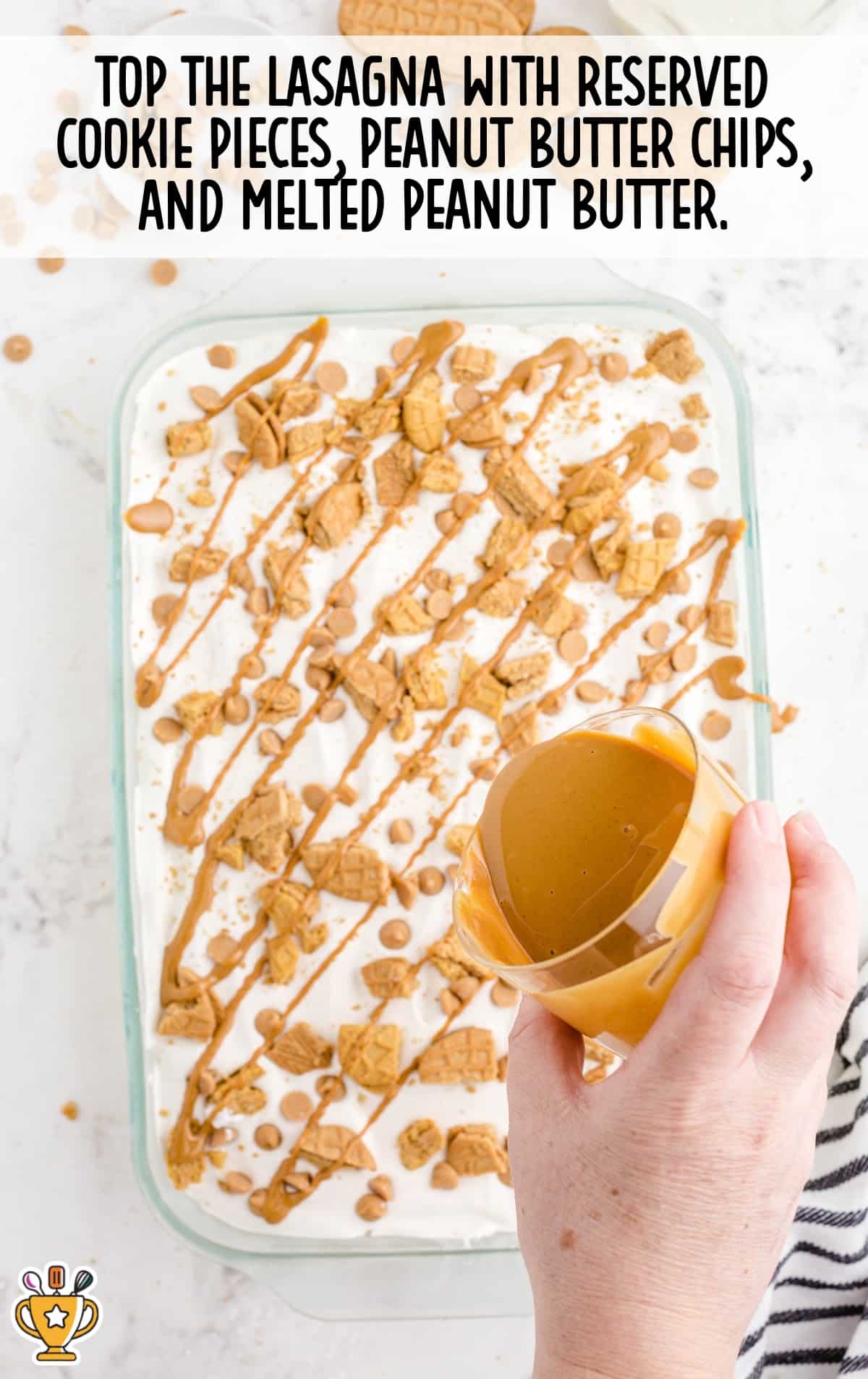 Overhead view of a hand drizzling melted peanut butter onto a dessert topped with whipped cream, crushed cookies, and peanut butter chips.