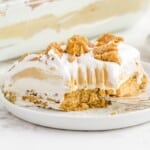 Slice of peanut butter lasagna on a white plate in front of a glass dish, highlighting its creamy layers and cookie base.
