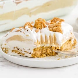 Slice of peanut butter lasagna on a white plate in front of a glass dish, highlighting its creamy layers and cookie base.