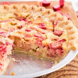Square image of a baked rhubarb custard pie in a metal pie tin, missing a large slice, with visible flaky crust and creamy filling.