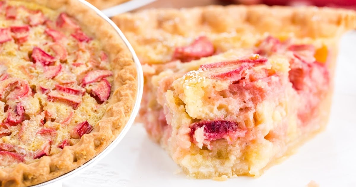 A slice of rhubarb custard pie placed in front of a full pie, both filled with pink rhubarb pieces and creamy custard.