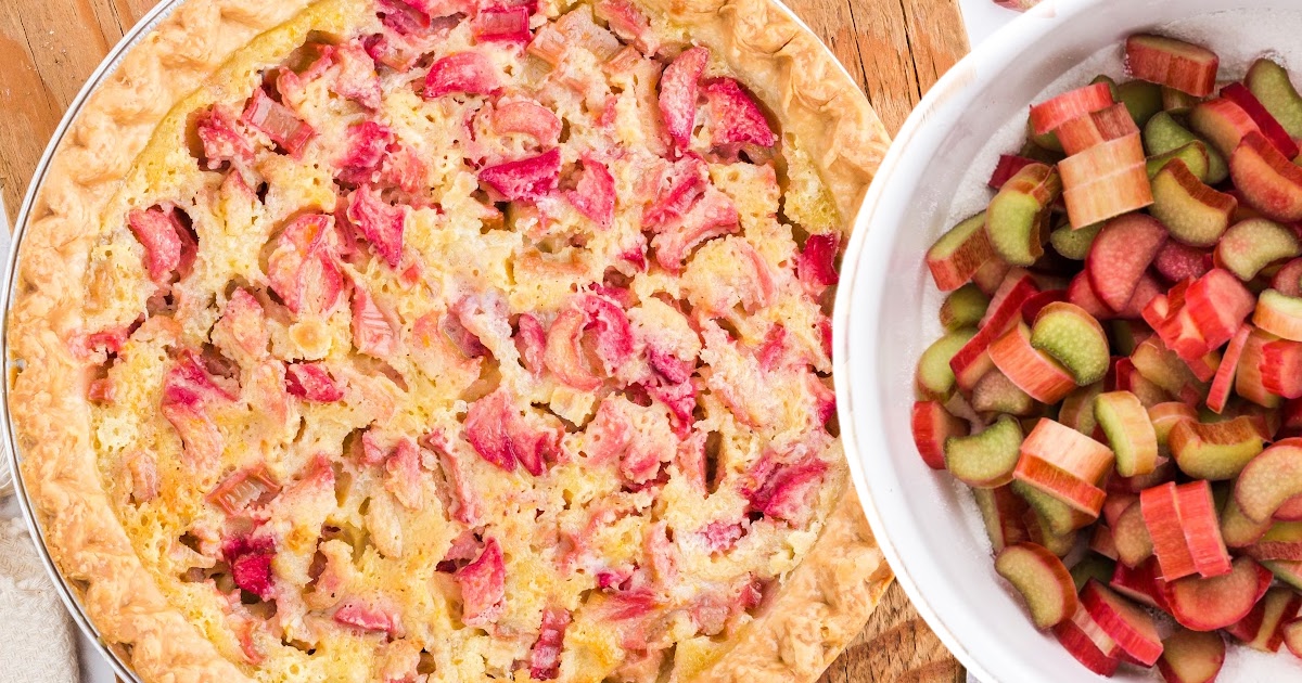 Top view of a rhubarb custard pie next to a white bowl filled with sliced raw rhubarb on a wooden surface.