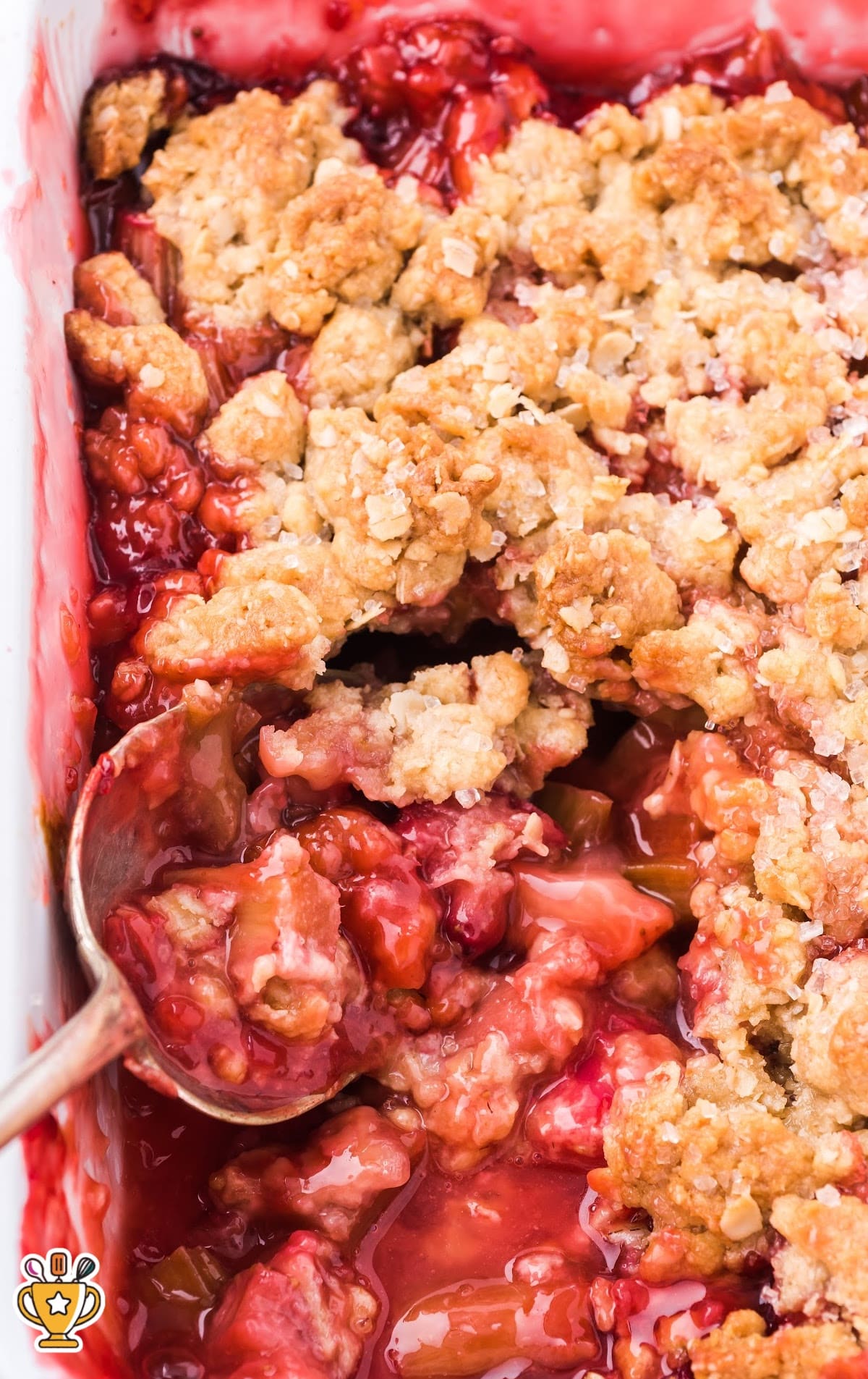 Close-up shot of a partially scooped rhubarb strawberry crisp with golden crumble and vibrant fruit filling in a white pan.