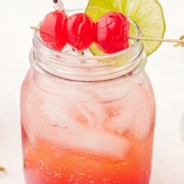 Front view of a Shirley Temple in a mason jar topped with three cherries on a skewer and lime slice, surrounded by grenadine, ginger ale, and cherries.