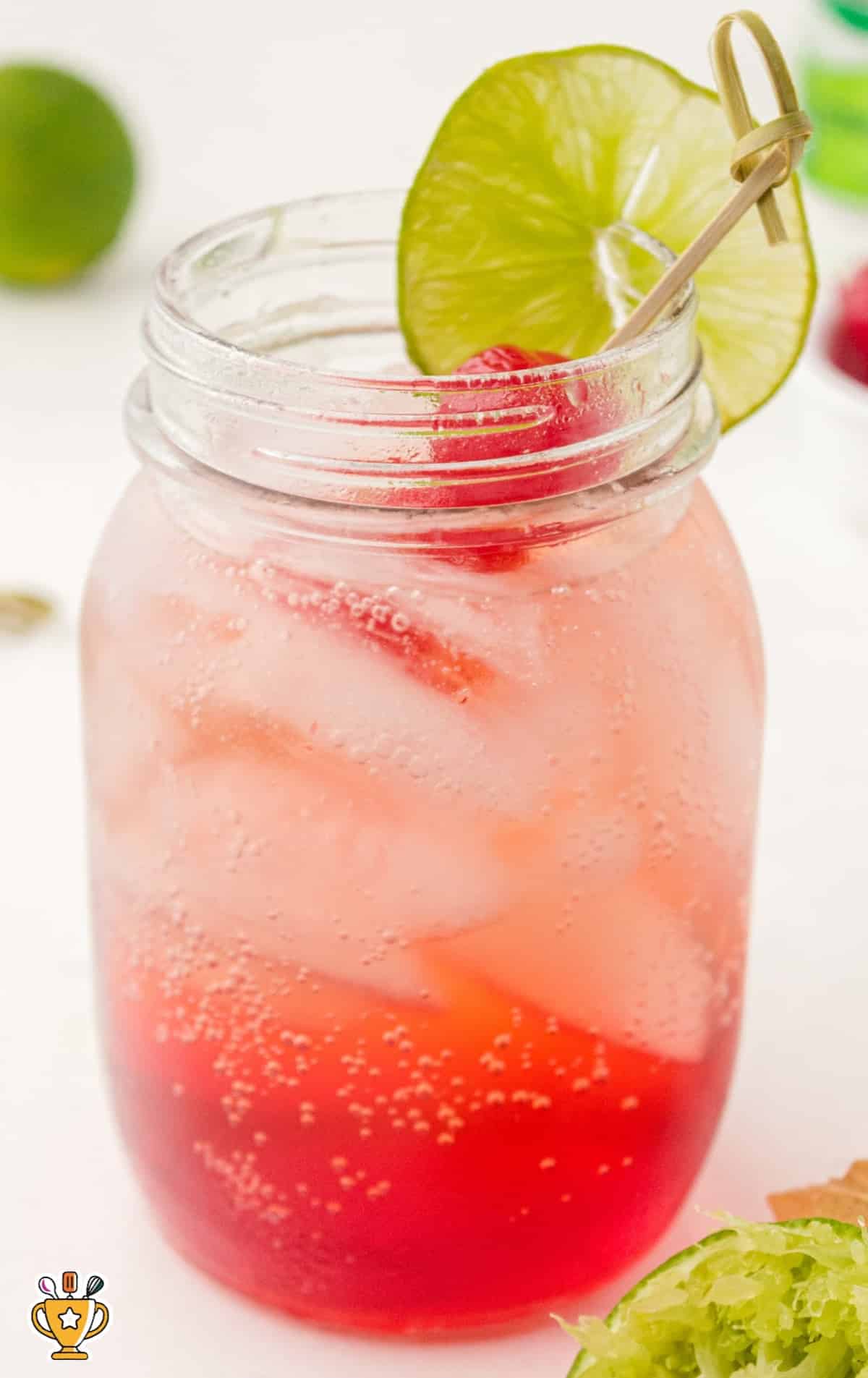 Classic Shirley Temple mocktail in a stemless wine glass with cherries and lime garnish, surrounded by drink ingredients in the background.