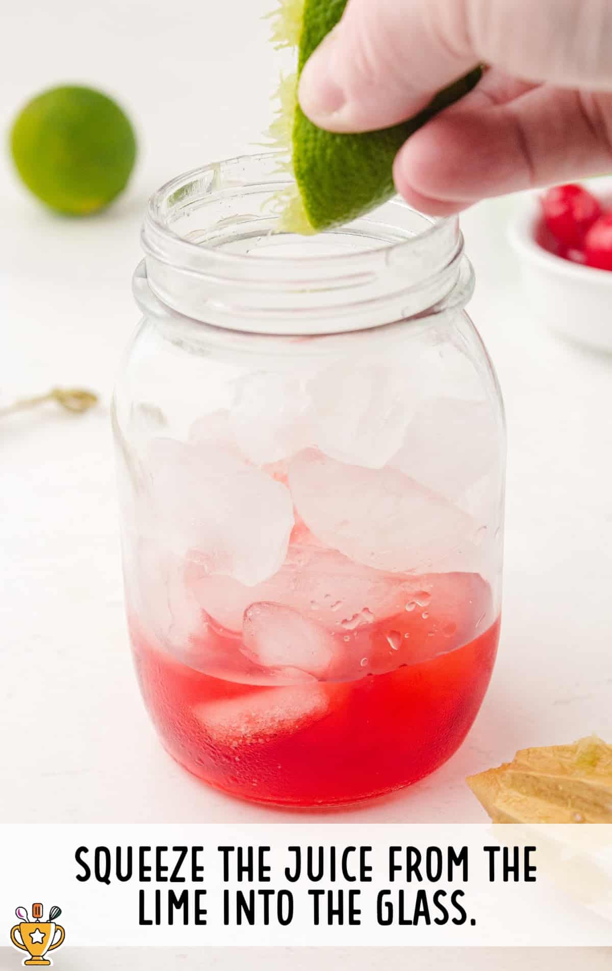 Hand squeezing fresh lime juice into a mason jar filled with ice and red grenadine syrup at the bottom of the glass.