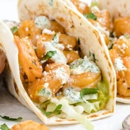 Close-up shot of shrimp tacos filled with juicy shrimp, shredded cabbage, guacamole, and cilantro lime crema on soft tortillas, with a bowl of creamy sauce and spoon nearby.