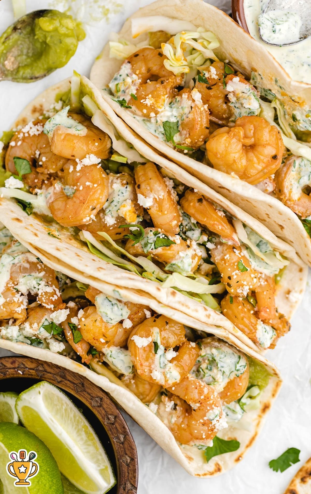 Top-down view of loaded shrimp tacos with vibrant toppings like guacamole, crema, cotija cheese, and cilantro, next to a small wooden bowl of lime wedges.