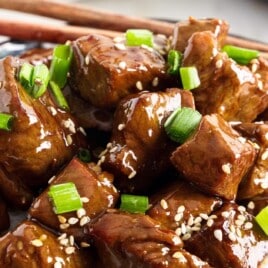 Close-up of glossy teriyaki steak bites garnished with sesame seeds and green onions in a ceramic bowl with chopsticks.