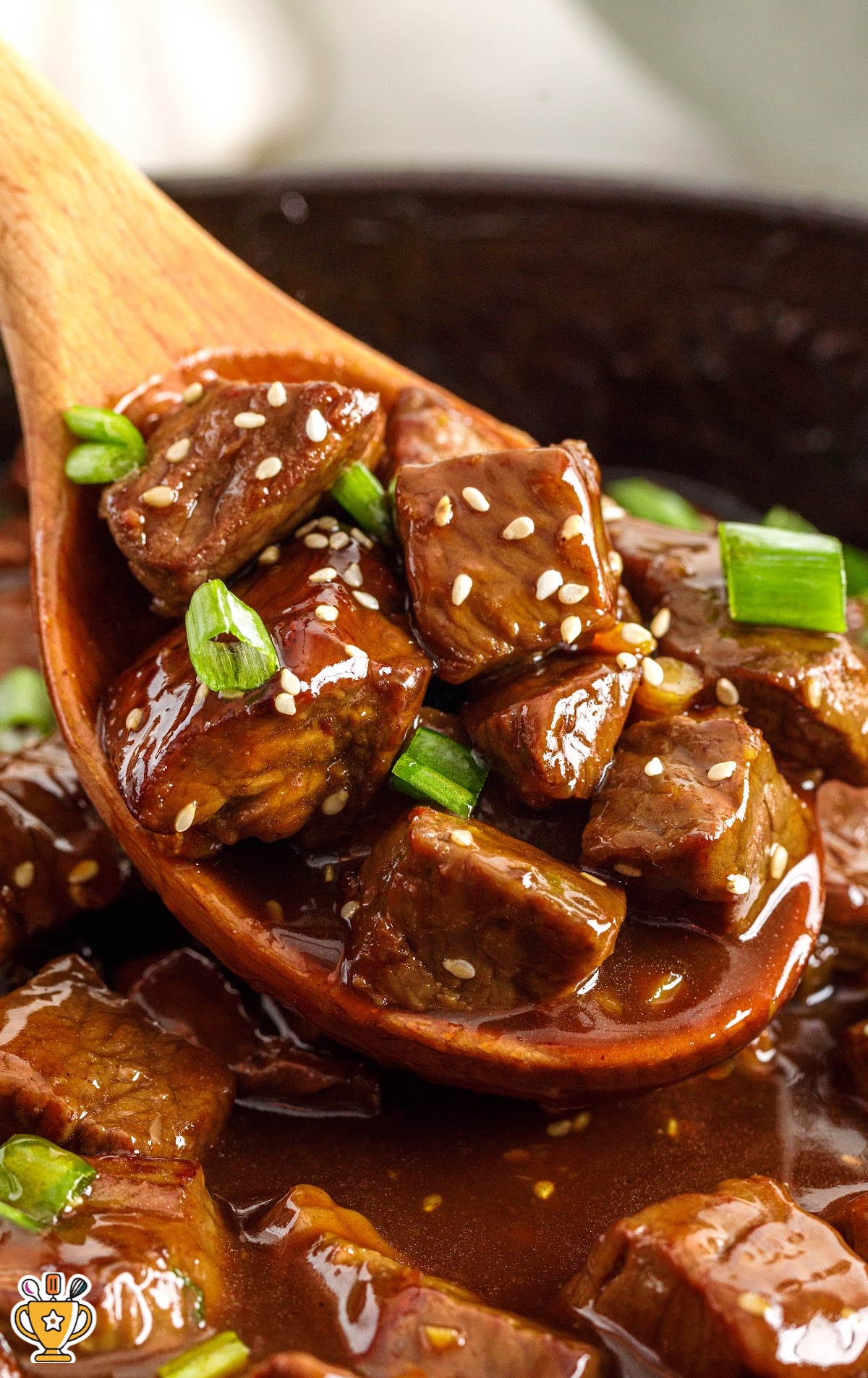 Wooden spoon lifting teriyaki steak bites from a pan, covered in thick sauce with sesame seeds and green onions.