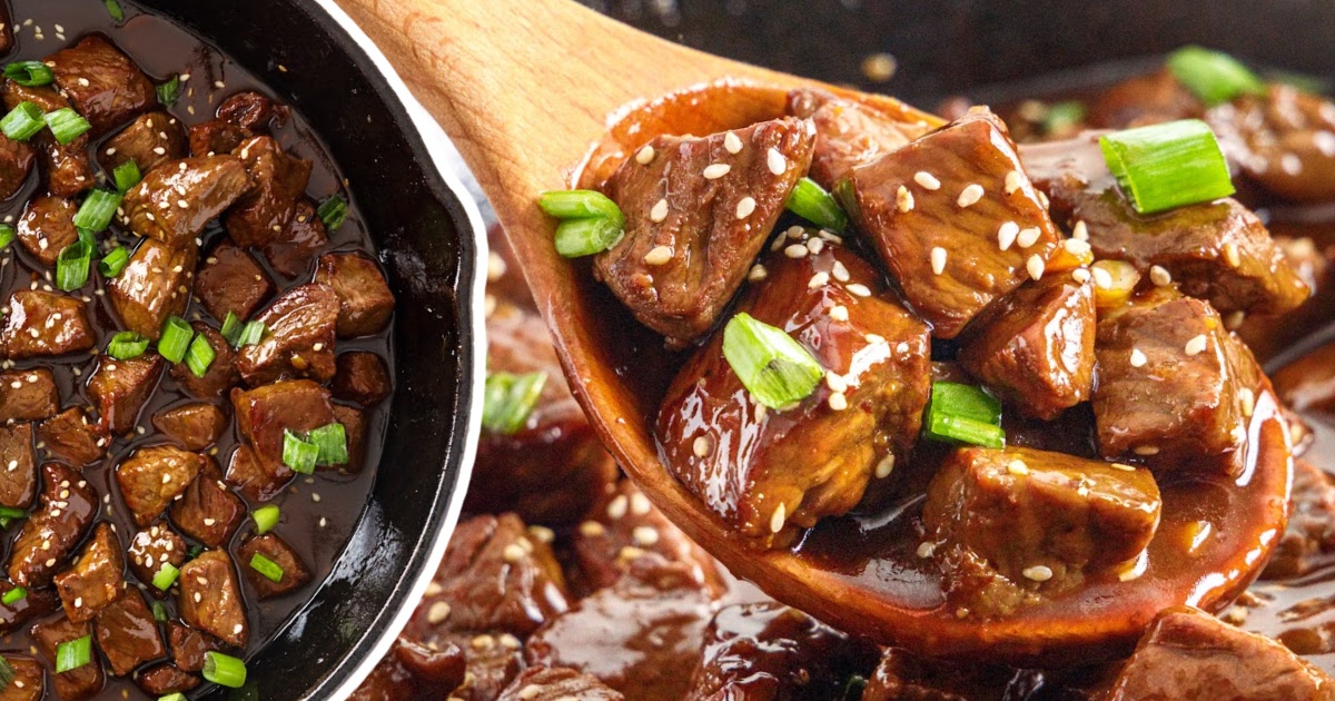 Teriyaki steak bites being scooped from a cast iron skillet with a wooden spoon, sauce glistening with garnishes.