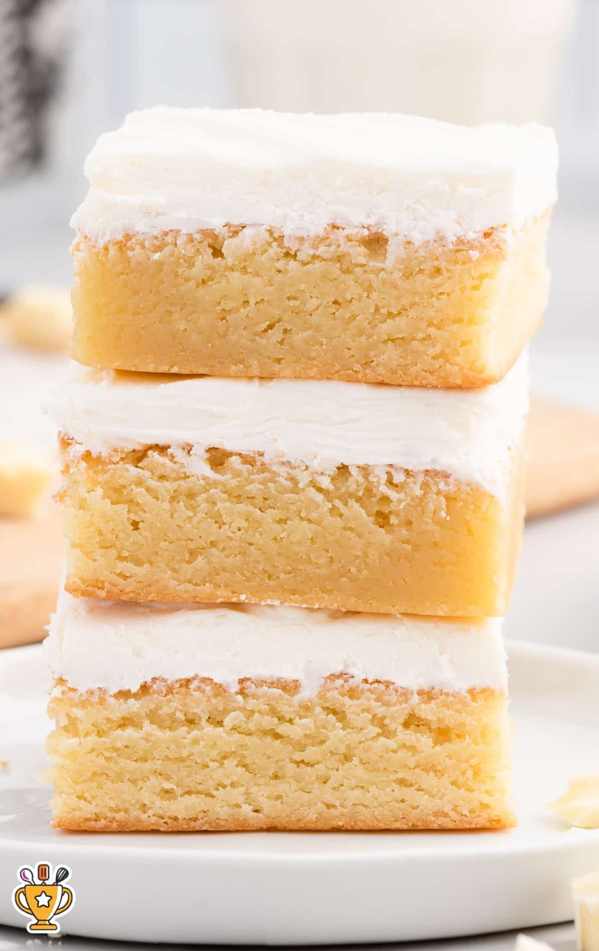 Three thick vanilla brownies stacked on a white plate, each topped with fluffy white frosting, perfect for dessert lovers.