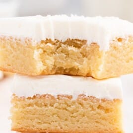 Two vanilla brownies stacked with a bite taken out of the top piece, showing a dense and gooey inside topped with creamy frosting.
