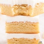 Three vanilla brownies stacked high on a white plate, each with a thick layer of white frosting and soft golden crumb texture.