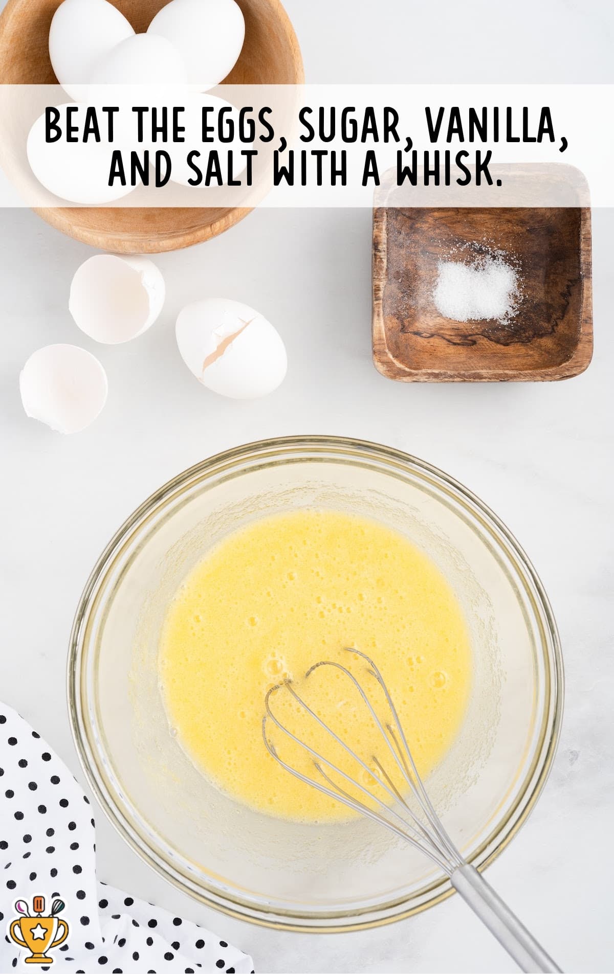 Overhead view of a mixing bowl filled with whisked eggs, sugar, vanilla, and salt, surrounded by cracked eggshells and salt on a white counter.