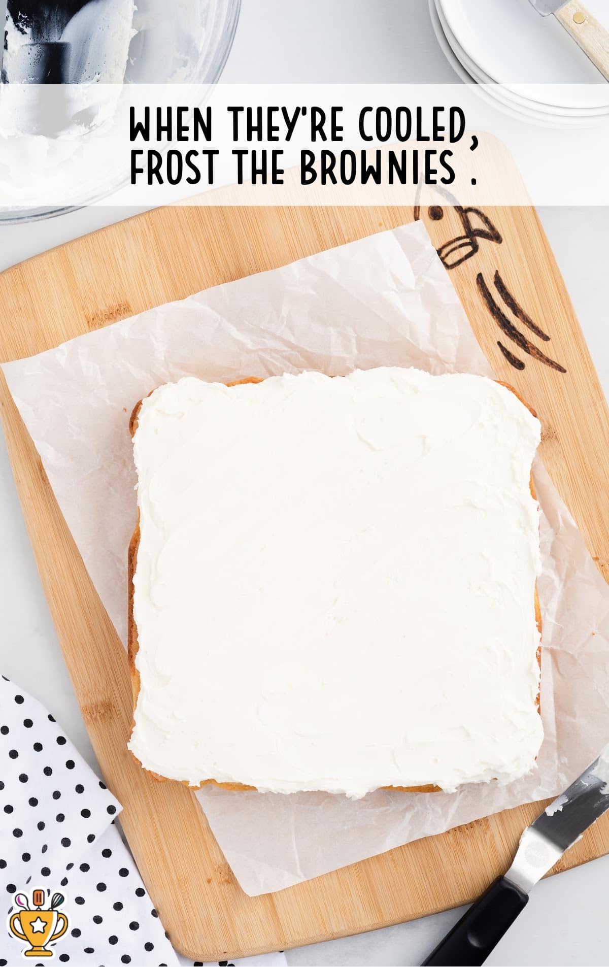 Fully frosted vanilla brownie slab resting on parchment and a wooden cutting board, ready to be sliced and served.
