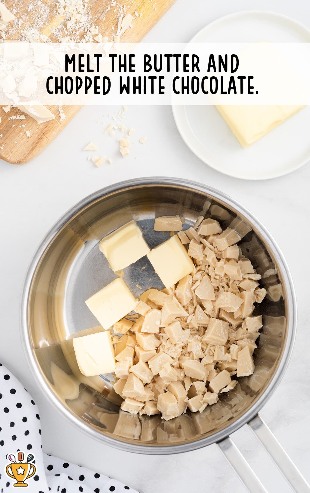 Butter and chopped white chocolate melting in a metal saucepan on a white background with a wooden cutting board and chocolate pieces nearby.