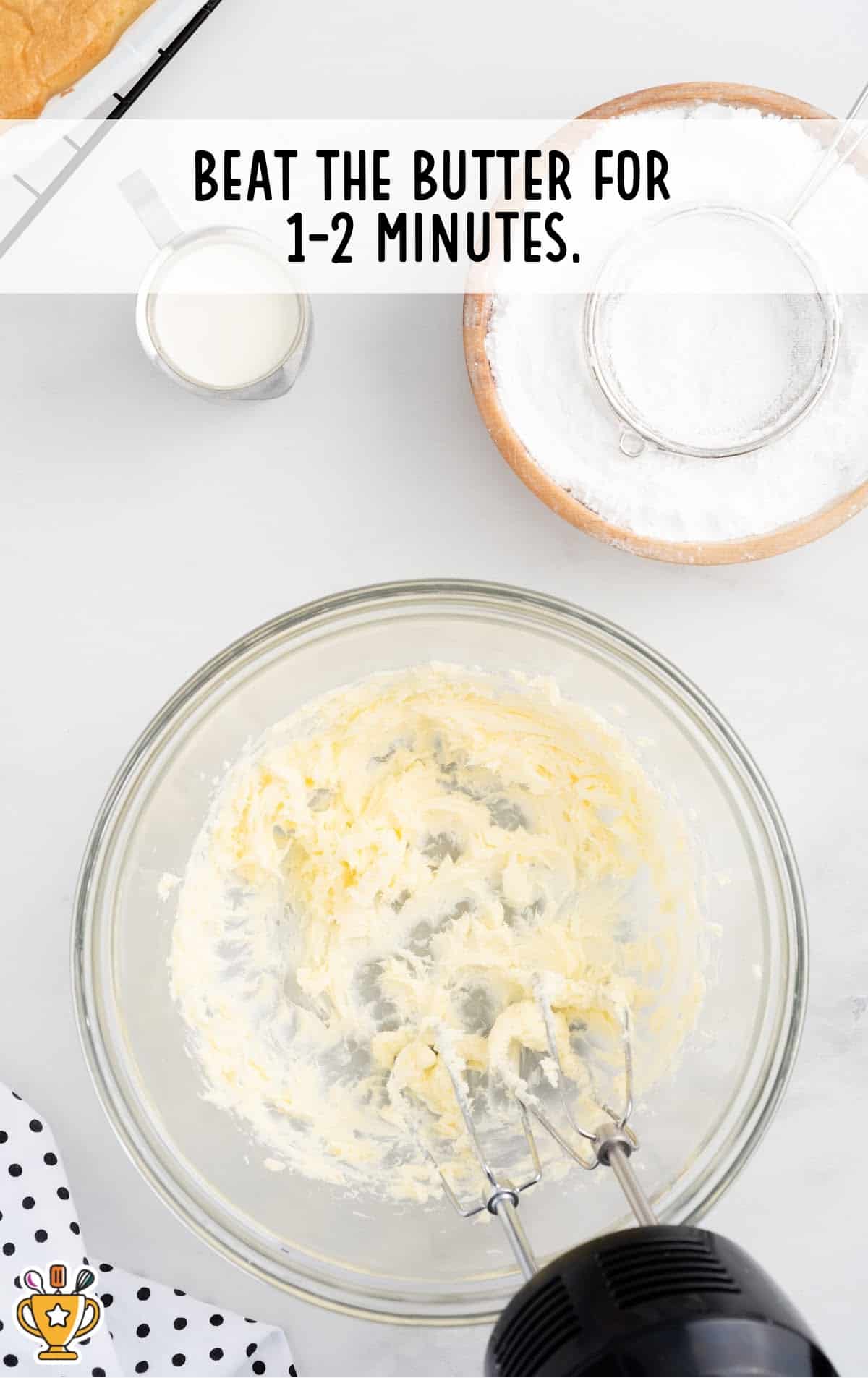Whipped butter in a glass bowl being prepared with a hand mixer, surrounded by powdered sugar and cream on a white kitchen surface.