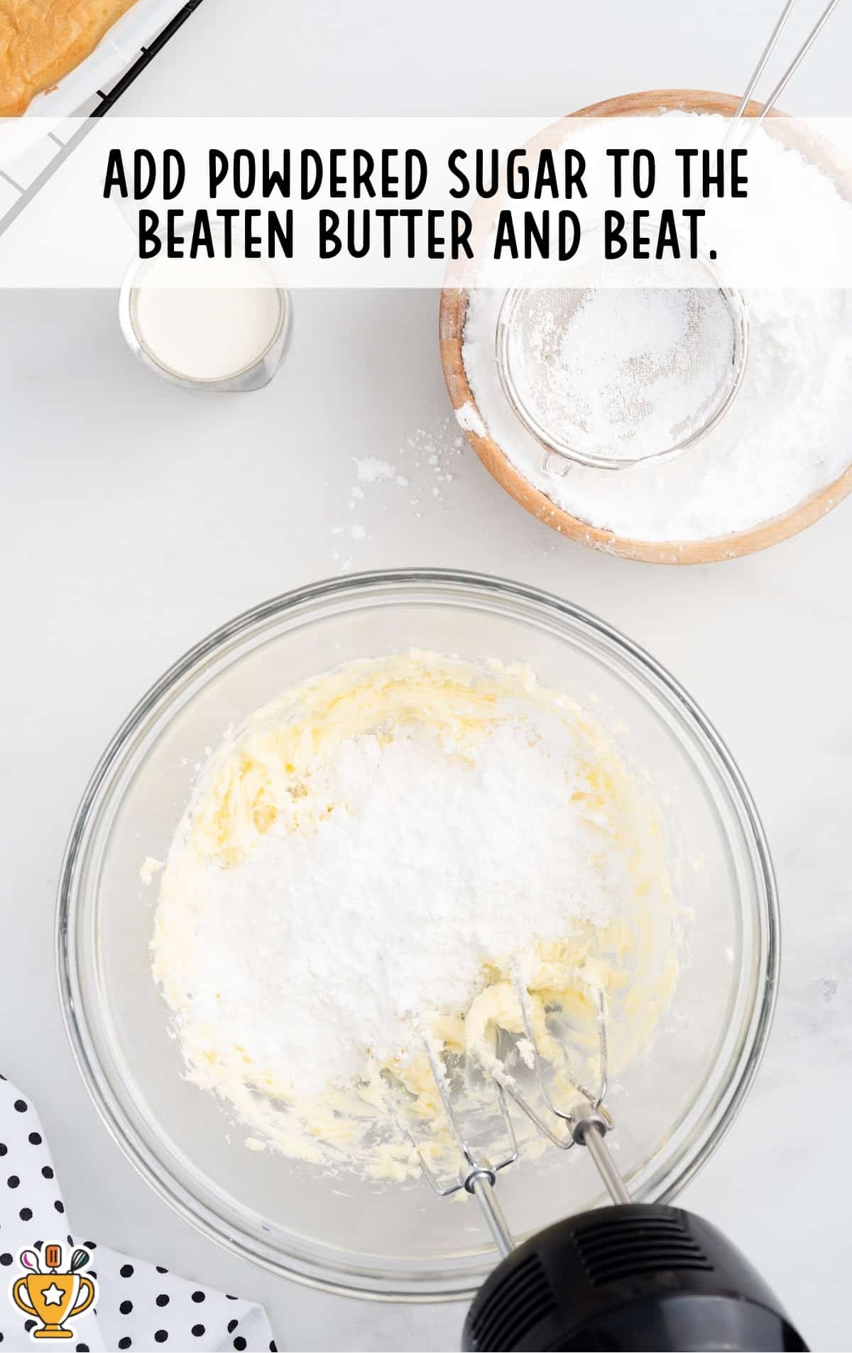 Electric mixer blending powdered sugar into beaten butter inside a glass mixing bowl, with extra ingredients staged around the workspace.