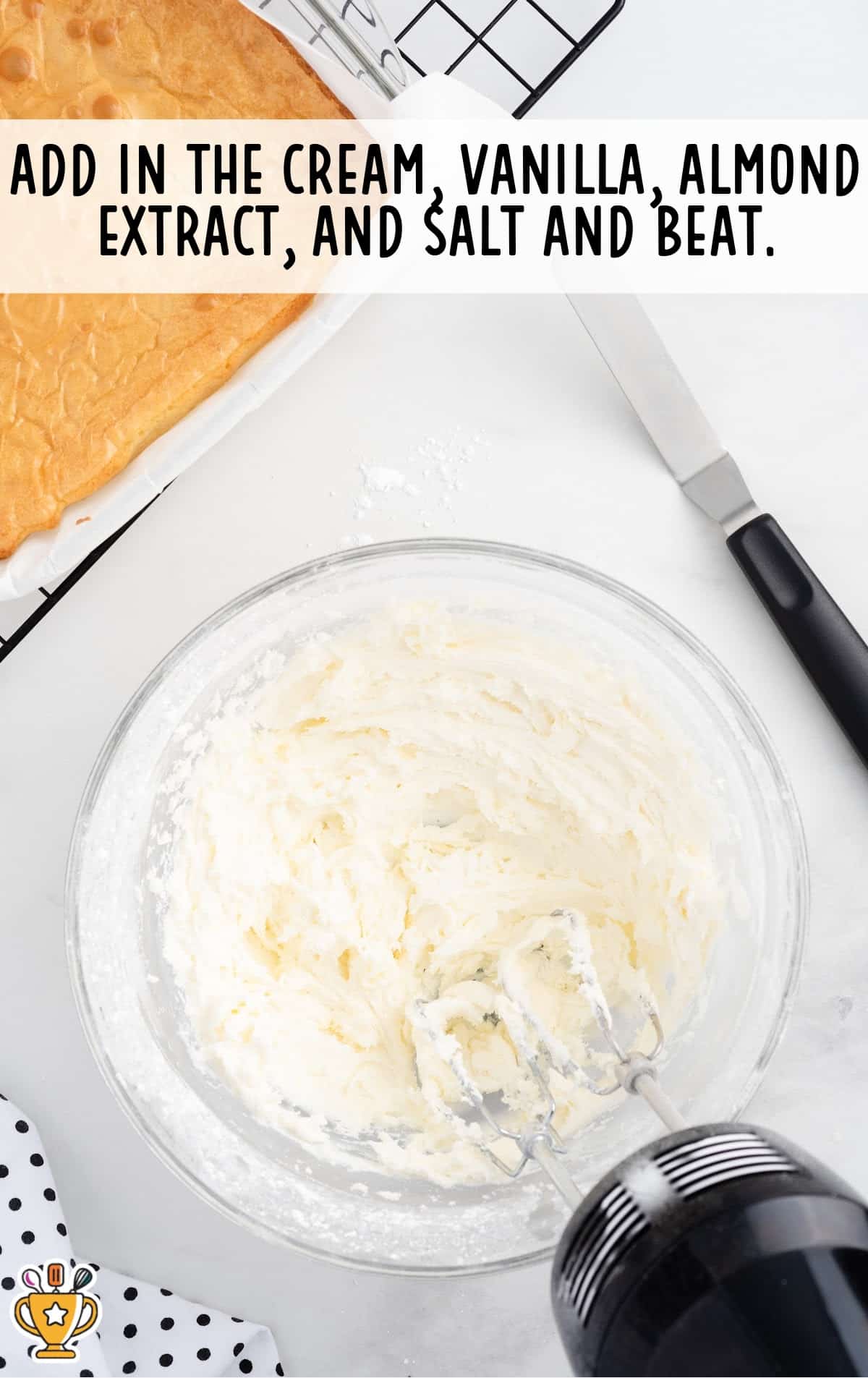 Creamy white frosting in a mixing bowl after adding cream, vanilla, almond extract, and salt, with ingredients and frosted brownies in the background.