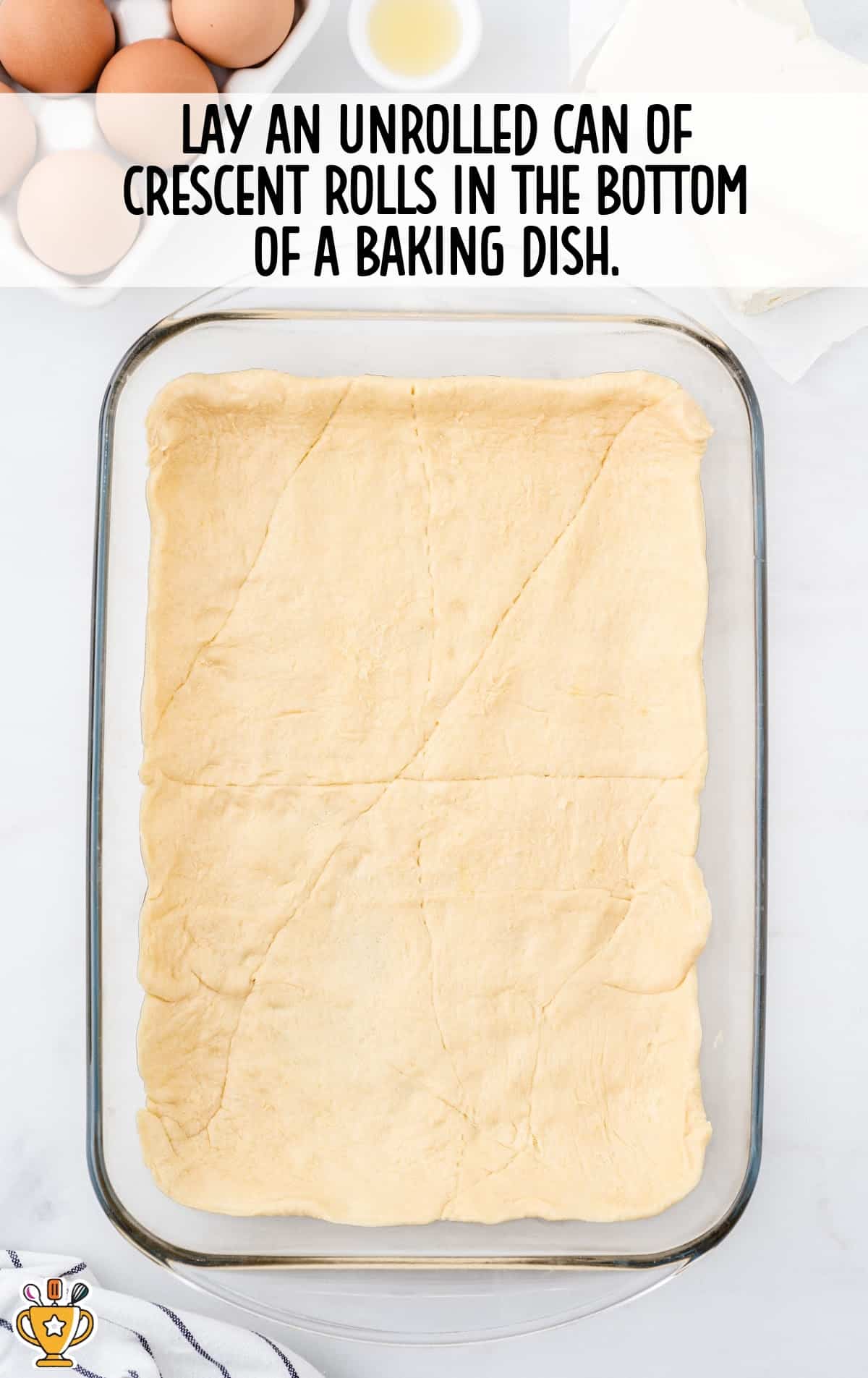 Unrolled crescent dough pressed into the bottom of a glass baking dish. Text overlay explains first step in recipe.