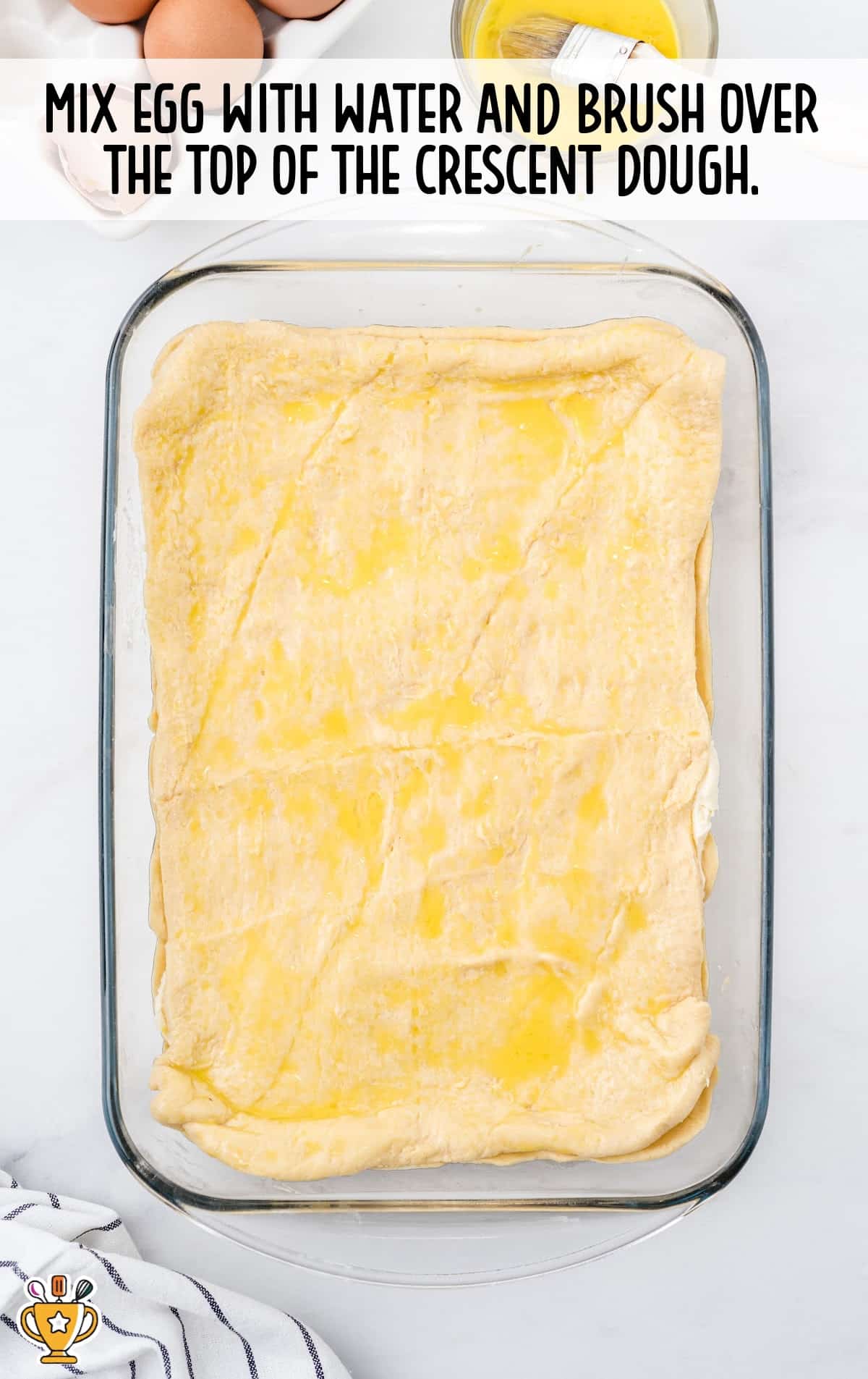 Top layer of crescent dough brushed with egg wash. Glass dish with dough edges crimped slightly. Text overlay shows baking step.